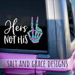 May include: A pink car with a white vinyl decal on the back window. The decal says "Hers Not His" and features a peace sign with a skeleton hand design. The peace sign is decorated with a colorful mosaic pattern.