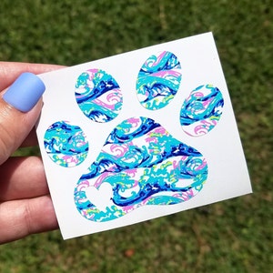 May include: A white adhesive decal featuring a paw print shape filled with a vibrant pattern of blue, turquoise, and pink swirling waves. The large central pad of the paw print includes a small, solid white heart cutout in the lower left corner.