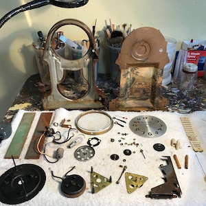 May include: Disassembled antique clock parts, including the clock face, hands, gears, and housing. The clock face is silver with black numerals. The housing has a landscape painting. Various tools and a magnifying lamp are in the background.