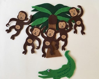 May include: Five brown felt monkeys hanging from a green felt palm tree with a brown felt trunk. A green felt alligator is lying on the ground below the tree.