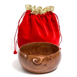 May include: A wooden yarn bowl with a carved spiral design and holes for yarn to pass through. The bowl is sitting on a white surface and is partially covered by a red velvet drawstring bag with gold trim.