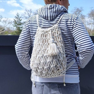 Könnte beinhalten: Ein weißer gehäkelter Rucksack mit einem Quasten. Der Rucksack wird von einer Person getragen, die ein gestreiftes Hemd und Jeans trägt.