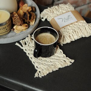 Może przedstawiać: Dwa białe podkładki pod kubki wykonane na szydełku z frędzlami, jeden z brązową etykietą z napisem "Nilloi Handmade with Love", oraz czarny kubek do kawy z kawą na czarnej powierzchni.