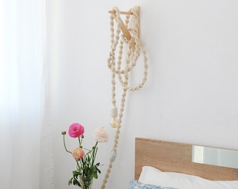 Op de afbeelding: Een witte touwlamp met een houten beugel, hangend boven een bed met een blauw-wit gepatroneerd kussen. De lamp is gemaakt van een dik, geknoopt touw en heeft een enkele gloeilamp. Het bed is van hout en heeft een wit hoofdeinde.
