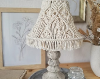 May include: A white macrame lampshade with a fringe trim sits on a gray concrete base with a wood grain pattern. The lamp is on a gray concrete tray with a white candle in a glass jar.