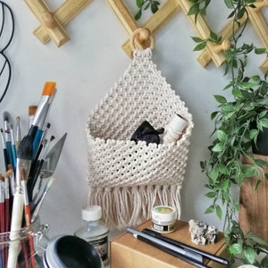 May include: A beige macrame wall hanging with a wooden ring at the top. The hanging has a pocket and a fringe at the bottom. It is hanging on a white wall with a wooden rack behind it.