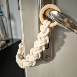 May include: A white rope door handle wrap with a wooden ring attached to a silver door handle.