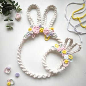 May include: Two white macrame bunny wall hangings with pastel pink, yellow, and green flower accents. The larger bunny is a full circle, while the smaller bunny is a half circle.
