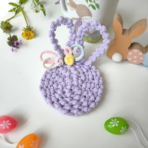 Könnte beinhalten: Ein lila gehäkelter Osterhase mit einer pastellrosa, gelben und blauen Schleife. Der Hase sitzt auf einer weißen Oberfläche.
