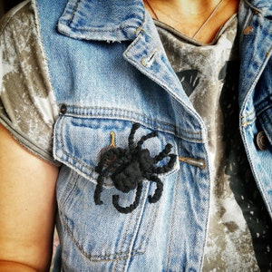 Könnte beinhalten: Eine schwarze gehäkelte Spinnennadel auf einer Jeansjacke. Die Spinne hat acht Beine und einen runden Körper.