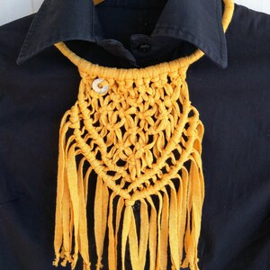 Può includere: Una collana in macramè gialla con una frangia e un piccolo bottone bianco. La collana è esposta su una camicia nera.