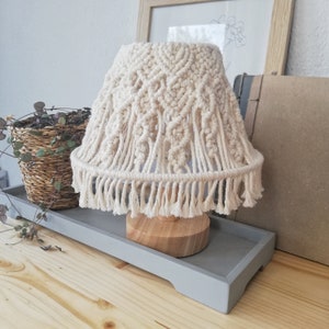 May include: A beige macrame lampshade with a fringe trim sits on a wooden base. The lampshade is on a grey tray on a wooden surface.