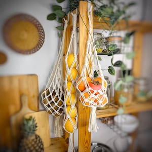 May include: Two white string bags hanging from a wooden rack, filled with oranges and persimmons. The bags are knotted at the top and have a fringe at the bottom.