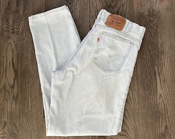 May include: Light-wash blue denim jeans featuring a high-waisted cut, a tan leather patch on the back waistband with Levi Strauss & Co. branding, and a small orange tab on the rear pocket. The jeans have a classic five-pocket design and straight legs.