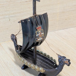 May include: A small black wooden model of a Viking ship with a sail and a dragon head prow. The ship has a gold trim and a crest on the sail that says "København".