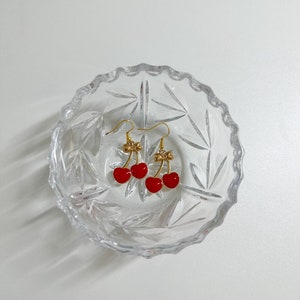 May include: A pair of gold cherry earrings with red glass cherries. The earrings are displayed in a clear glass bowl.