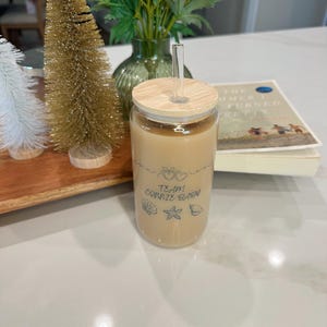 May include: A clear glass tumbler filled with a light brown beverage, featuring a wooden lid and a clear straw. The tumbler has the text "Team Connie Brew" with heart, seashell, starfish, and shell graphics. A book and decorative trees are in the background.