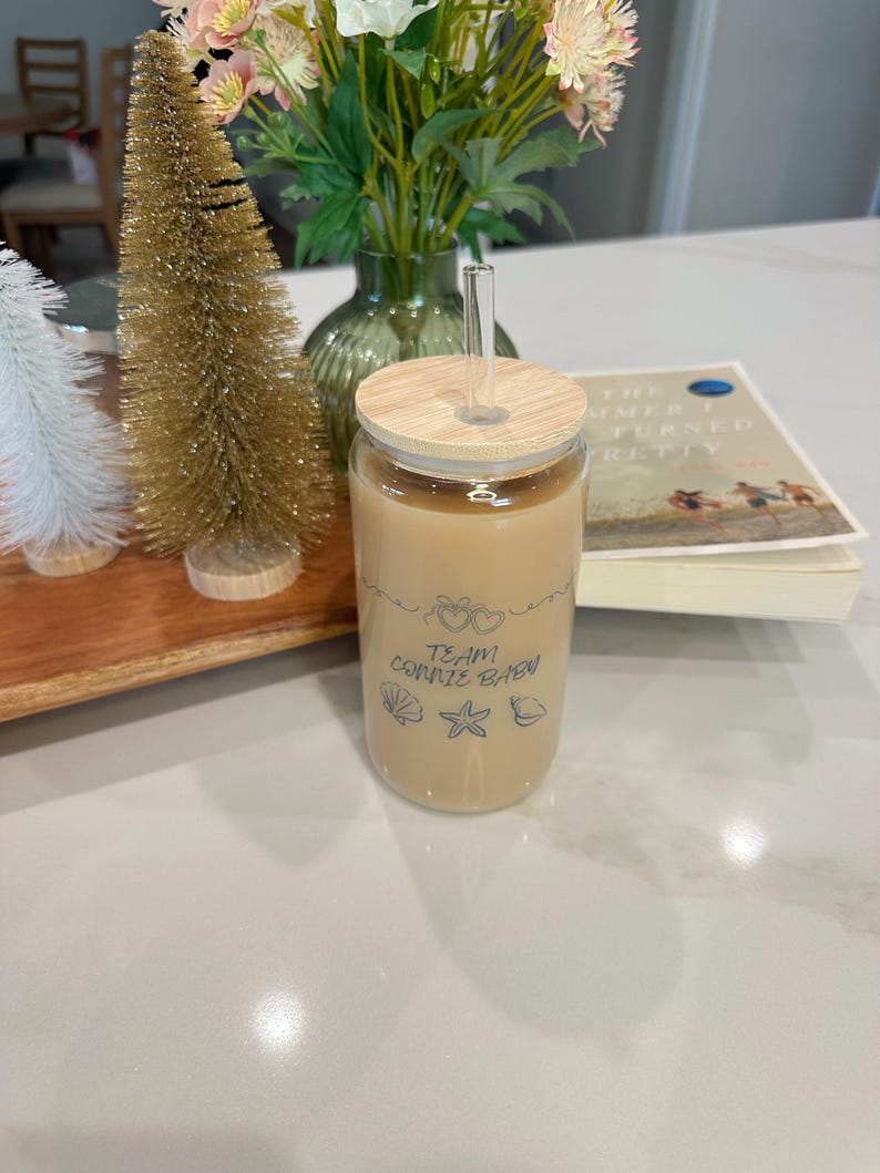 May include: A clear glass tumbler filled with a light brown beverage, featuring a wooden lid and a clear straw. The tumbler has the text "Team Connie Brew" with heart, seashell, starfish, and shell graphics. A book and decorative trees are in the background.