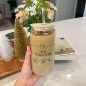 May include: A clear glass tumbler filled with a light brown beverage, featuring a bamboo lid and a reusable straw. The tumbler is decorated with the text "TEAM CONNIE BABY" and seashell graphics. The drink is held by a person.