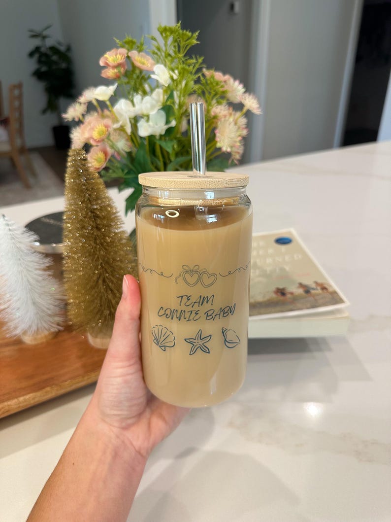 May include: A clear glass tumbler filled with a light brown beverage, featuring a bamboo lid and a reusable straw. The tumbler is decorated with the text "TEAM CONNIE BABY" and seashell graphics. The drink is held by a person.