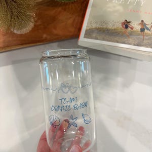 May include: Clear glass can-shaped drinking glass with blue text and illustrations. The text reads "TEAM CONNIE BABY" with heart, seashell, starfish, and conch shell illustrations. The glass is held by a person.
