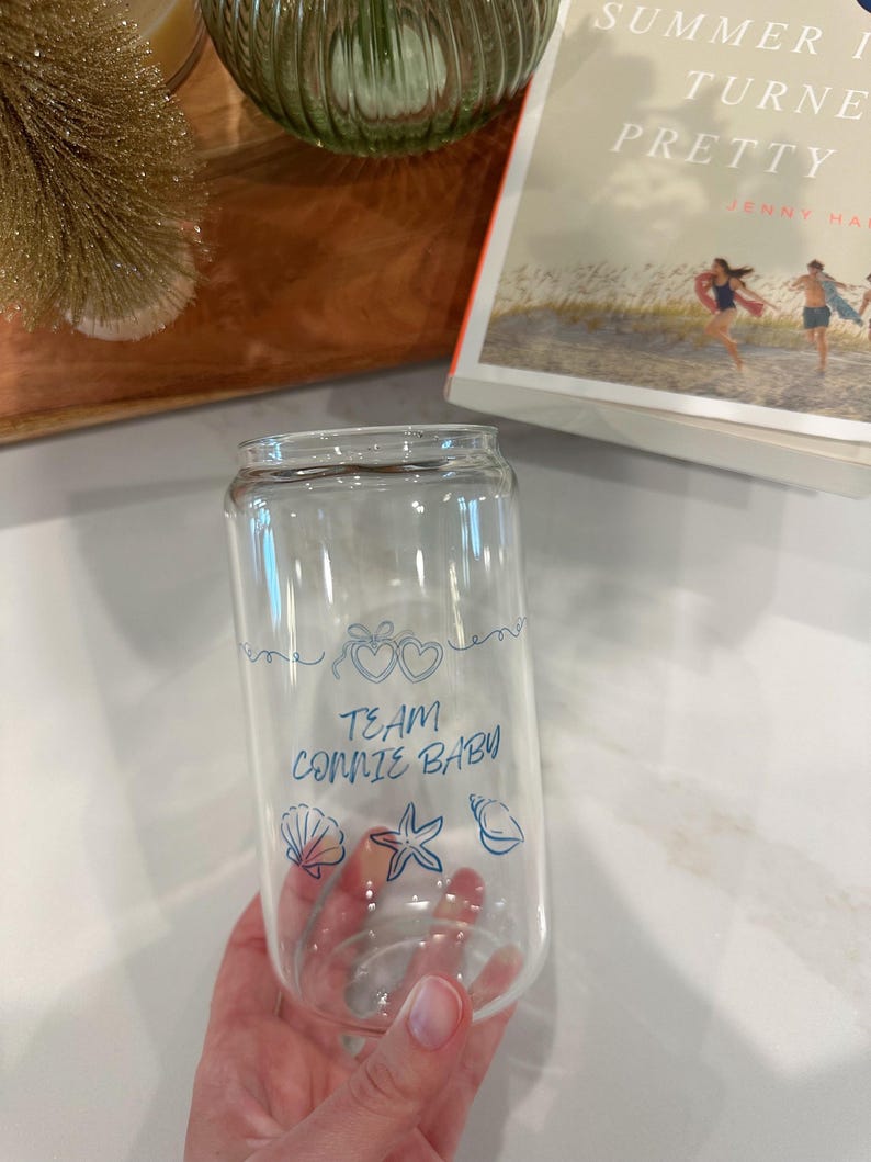 May include: Clear glass can-shaped drinking glass with blue text and illustrations. The text reads "TEAM CONNIE BABY" with heart, seashell, starfish, and conch shell illustrations. The glass is held by a person.