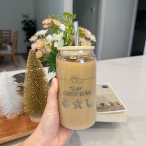 May include: A clear glass tumbler filled with a light brown beverage, featuring a bamboo lid and a silver straw. The glass is decorated with the text "TEAM CONNIE BABY" and seashell graphics. The tumbler is held in a hand.