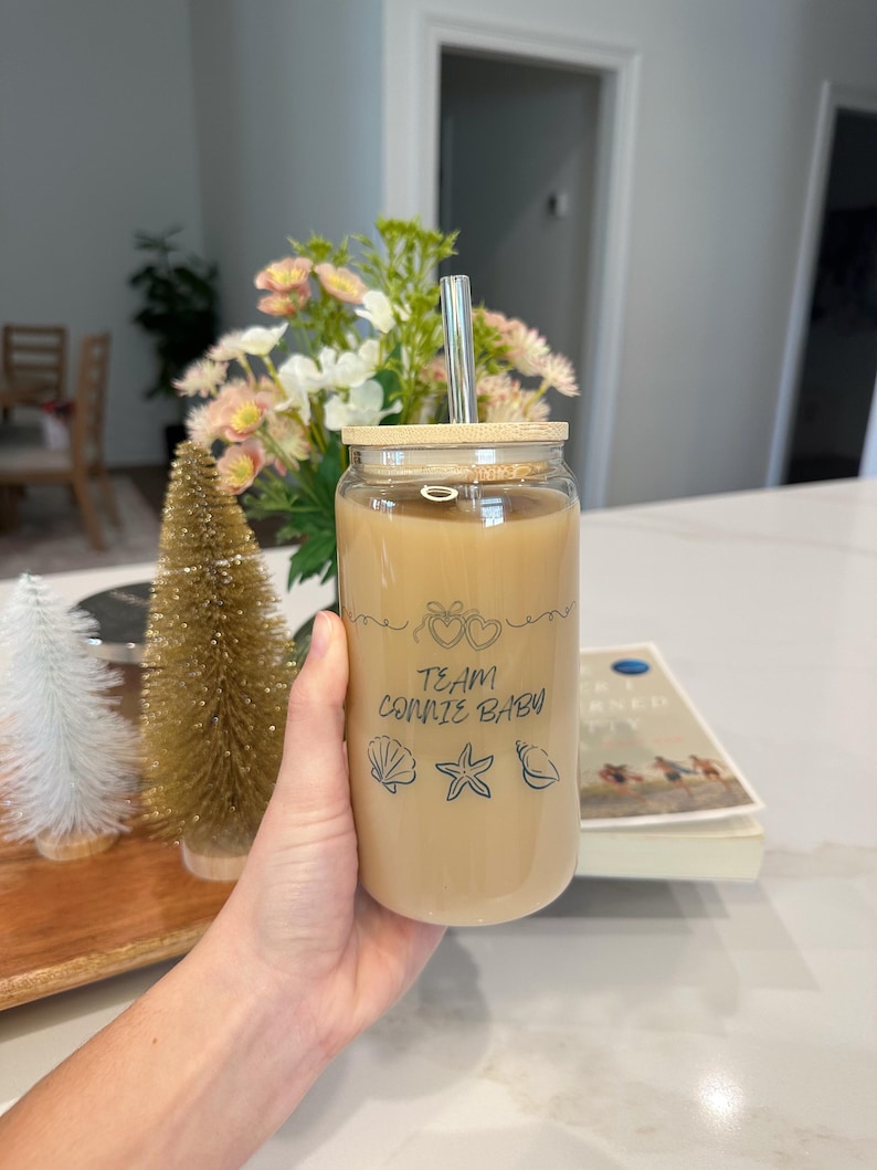 May include: A clear glass tumbler filled with a light brown beverage, featuring a bamboo lid and a silver straw. The glass is decorated with the text "TEAM CONNIE BABY" and seashell graphics. The tumbler is held in a hand.