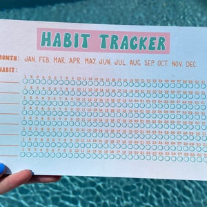 Può includere: Un tracker delle abitudini stampato su carta bianca con sfondo azzurro chiaro. Il titolo "HABIT TRACKER" è in rosa e verde. Il tracker include mesi, date e spazi per monitorare le abitudini. La carta è tenuta da una persona.