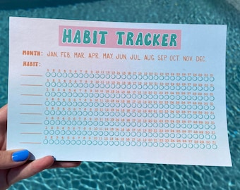May include: A habit tracker printed on a white paper with a light blue background. The title "HABIT TRACKER" is in pink and green. The tracker includes months, dates, and spaces to track habits. The paper is held by a person.