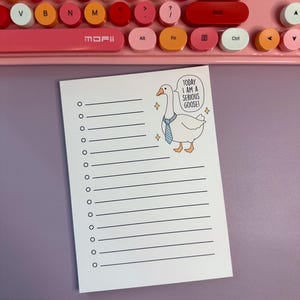 Op de afbeelding: Een wit notitieblok met een gelijnde lay-out en een cartoonillustratie van een gans met een blauwe stropdas. De gans heeft een tekstballon met de tekst "VANDAAG BEN IK EEN SERIEUZE GANS!" Op de achtergrond staat een roze en wit toetsenbord.