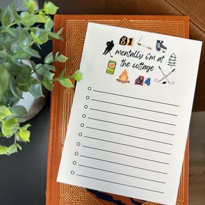 Pode incluir: Um bloco de notas branco com a frase "mentally I'm at the cottage" e ilustrações temáticas de cabanas. O bloco de notas está em cima de um livro castanho com padrões decorativos dourados. O bloco de notas tem uma lista de linhas em branco.