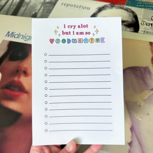 May include: A white notepad with the text "i cry a lot but I am so productive" in colorful, playful lettering. The notepad has a series of lined spaces for list-making. The background includes album covers.