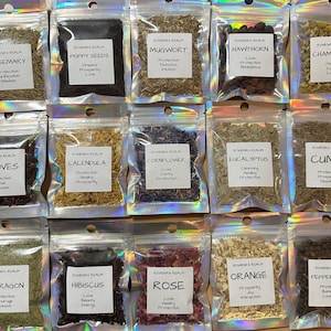 May include: A collection of small, clear, resealable bags filled with various dried herbs and seeds. Each bag has a white label with the name of the herb and associated properties, such as "Rose" for love and protection. The bags are arranged in neat rows.