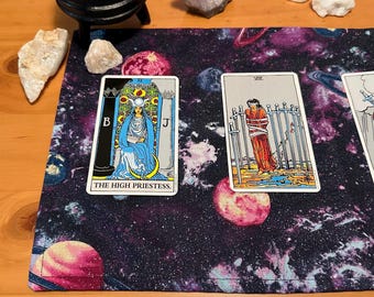 May include: A tarot card reading setup featuring a purple and black galaxy-themed mat. Two tarot cards are visible: "The High Priestess" and another with a figure bound by swords. Crystals and a small cauldron are also present.