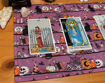 May include: A purple tarot cloth with a Halloween theme featuring black cats, skulls, pumpkins, and potion bottles. Two tarot cards are displayed: "Eight of Swords" and "The High Priestess". The cloth is on a wooden surface with crystals and a black cauldron.
