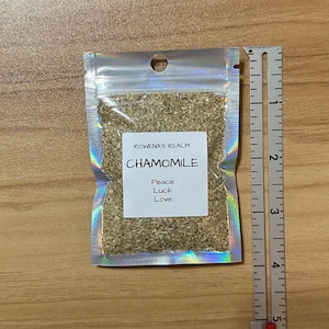 May include: A small, clear, iridescent bag filled with dried chamomile flowers. The bag has a label that reads "ROWENA'S REALM CHAMOMILE Peace Luck Love". A ruler is next to the bag, showing measurements in inches.