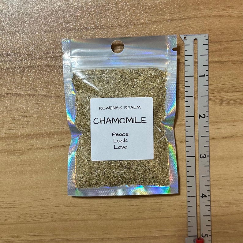 May include: A small, clear, iridescent bag filled with dried chamomile flowers. The bag has a label that reads "ROWENA'S REALM CHAMOMILE Peace Luck Love". A ruler is next to the bag, showing measurements in inches.
