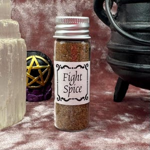 May include: A small glass bottle with a silver lid filled with brown spice. The bottle has a white label with black text that reads "Fight Spice".