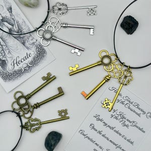 May include: Two black cord necklaces featuring sets of metal skeleton keys in silver, gold, and bronze tones. The keys have intricate bow designs and are displayed alongside two printed cards, one depicting Hecate and the other with text.