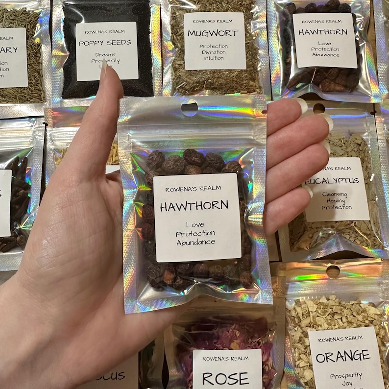 May include: A hand holding a small, clear, metallic pouch of Hawthorn berries. The pouch has a white label with the text "HAWTHORN, Love, Protection, Abundance." Other pouches with herbs and labels are in the background.