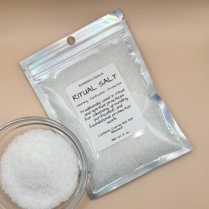 May include: A clear glass bowl filled with white coarse sea salt next to a sealed, clear bag of ritual salt. The bag has a label that reads "RITUAL SALT" and "Contents Coarse Sea Salt (Blessed)". The net weight is 5 oz.