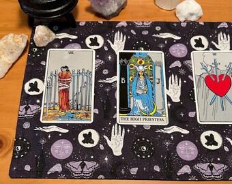 May include: A tarot card reading setup featuring three cards on a dark purple mat with white hand and moth patterns. The cards include "The High Priestess" and two other cards. Crystals and a cauldron are in the background.