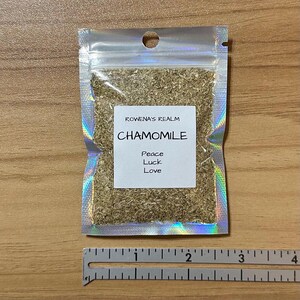 May include: A small, clear, iridescent pouch filled with dried chamomile flowers. The pouch has a white label with the text "ROWENA'S REALM CHAMOMILE Peace Luck Love". A ruler is at the bottom of the image.