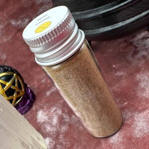 K&ouml;nnte beinhalten: Ein kleines Glasgef&auml;&szlig; mit einem silbernen Deckel, gef&uuml;llt mit braunem Pulver. Der Deckel hat einen gelben Aufkleber mit dem Text "Sorcery" und "15ml".