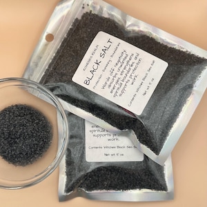 May include: Two sealed pouches of black salt, labeled "Black Salt" with text describing its uses. A small glass bowl contains a portion of the black salt. The labels state the contents are Witches Black Sea Salt, net weight 5 oz.