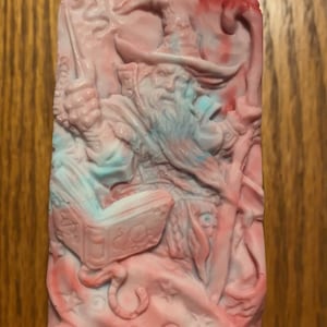 May include: A pink and blue soap bar featuring a detailed design of a wizard holding a book and a staff. The wizard has a long beard and is wearing a hat.