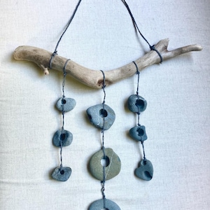 Puede incluir: Un colgante de pared rústico con una rama de madera flotante natural y varias piedras grises suspendidas con hilo oscuro. Las piedras varían en tamaño y forma, algunas con agujeros, creando un diseño único y orgánico. Los tonos neutros crean una estética relajante.