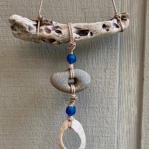 Puede incluir: Móvil artesanal de madera flotante con una rama clara y desgastada. Debajo, una piedra gris con un agujero está suspendida con hilo, junto con cuentas de vidrio azul y un colgante blanco en forma de hueso. Fondo de madera verde claro.