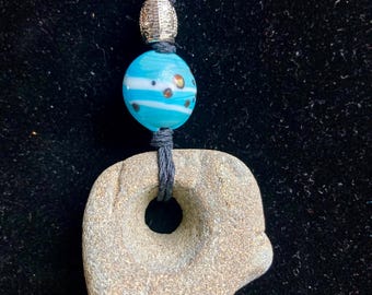 May include: A handmade necklace featuring a natural stone pendant with a hole, a blue and white glass bead, and a silver-colored bead. The necklace is strung on a dark gray cord, creating a unique and earthy accessory.